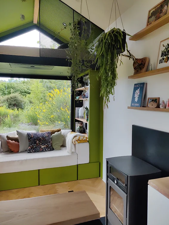 Aménagement intérieur d'une tiny house. Espace canapé dans niche verte, poêle à bois et étagères en chêne.