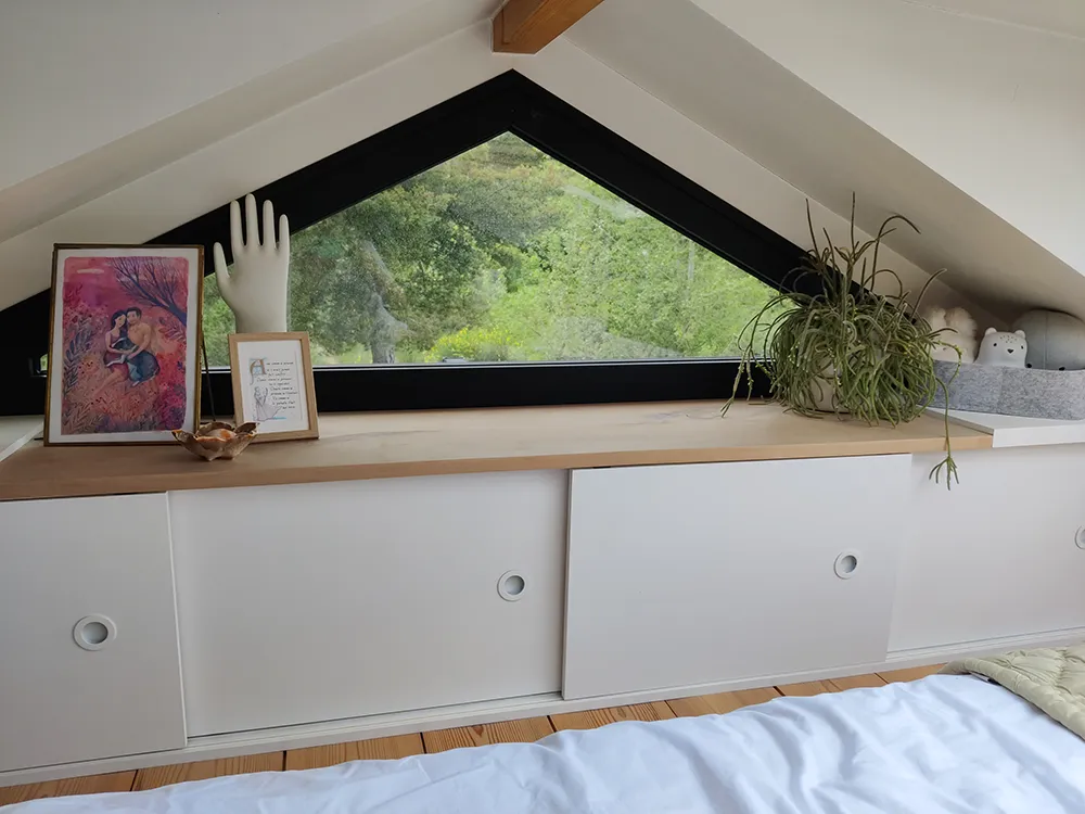 Aménagement intérieur d'une tiny house. Meuble de rangement à portes coulissantes pour vêtement et fenêtre triangulaire vers nature alentours.