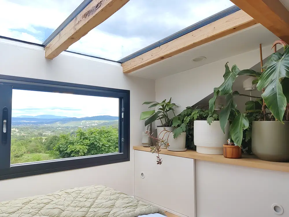 Aménagement intérieur d'une tiny house. Fenêtre de toit au-dessus du lit double et rangements longs à vêtements avec plantes vertes.