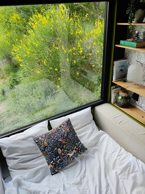 Aménagement intérieur d'une tiny house. Canapé convertible lit double avec vue dehors sur les genêts.