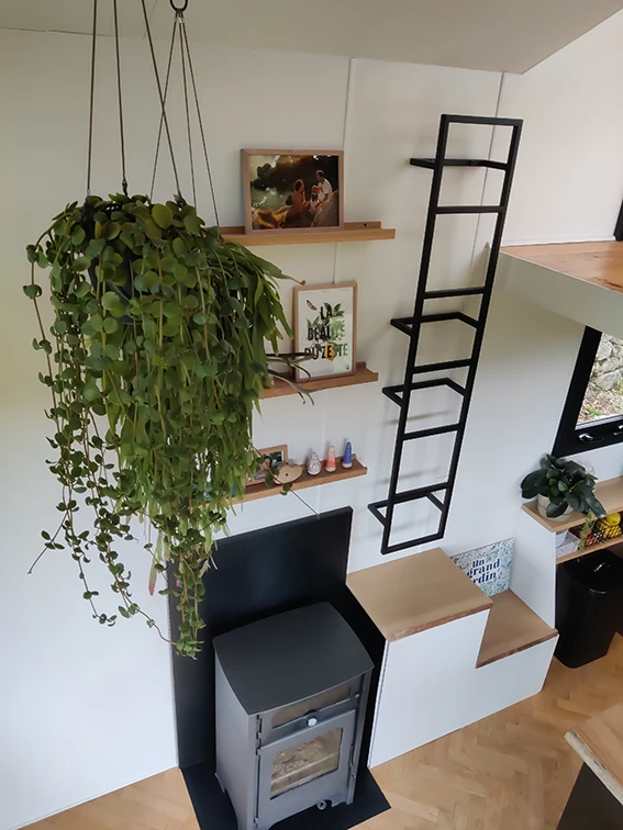 Aménagement intérieur d'une tiny house. Vue de haut de l'échelle vers mezzanine double et poêle à bois avec étagères et plantes suspendues.